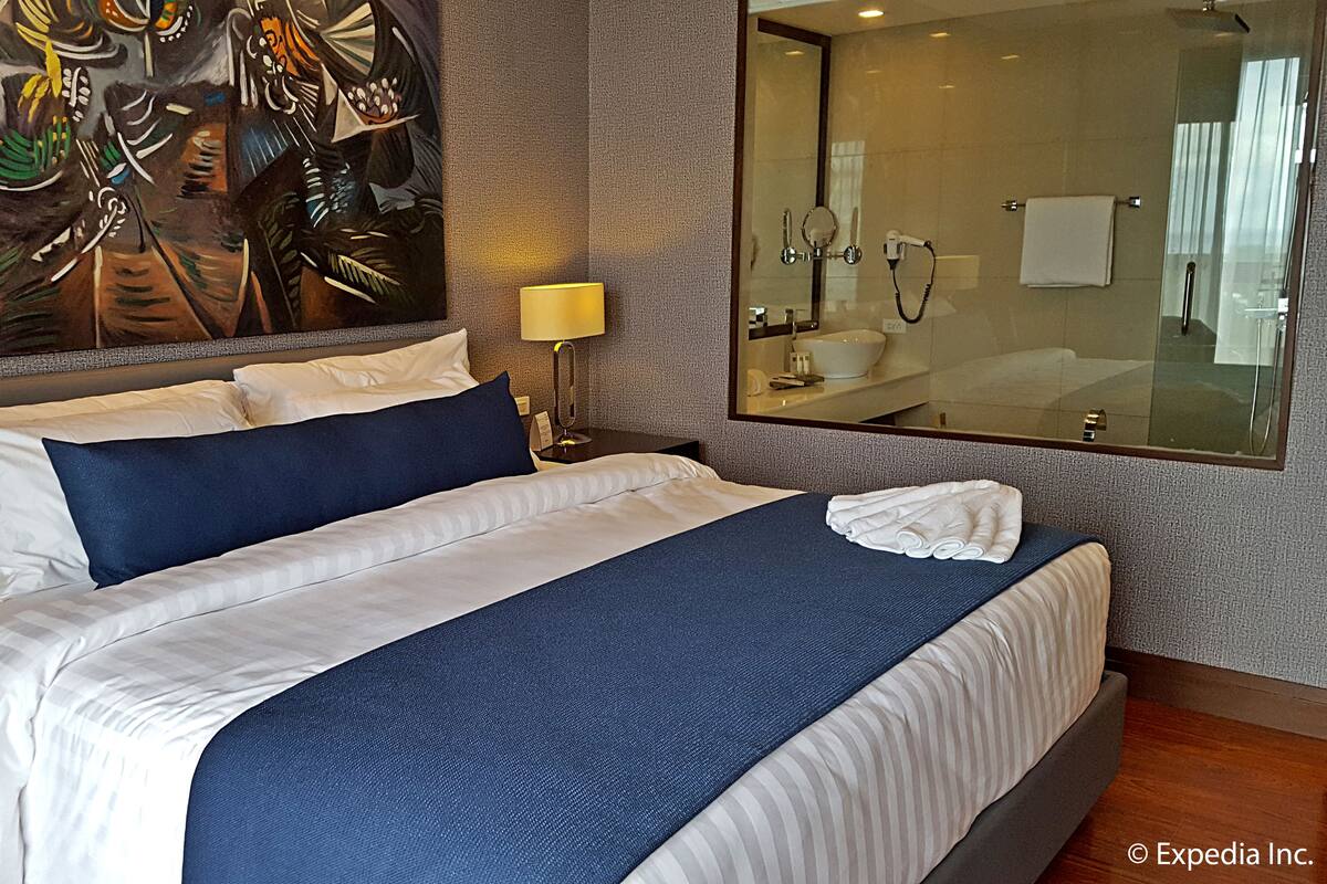 Une chambre d’hôtel avec un grand lit, une table de chevet avec un téléphone, une lampe fixée au mur et un miroir au-dessus du lavabo.