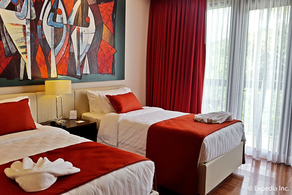 Une chambre d’hôtel avec deux lits, une literie rouge et blanche, un tableau au mur et une fenêtre avec des rideaux.
