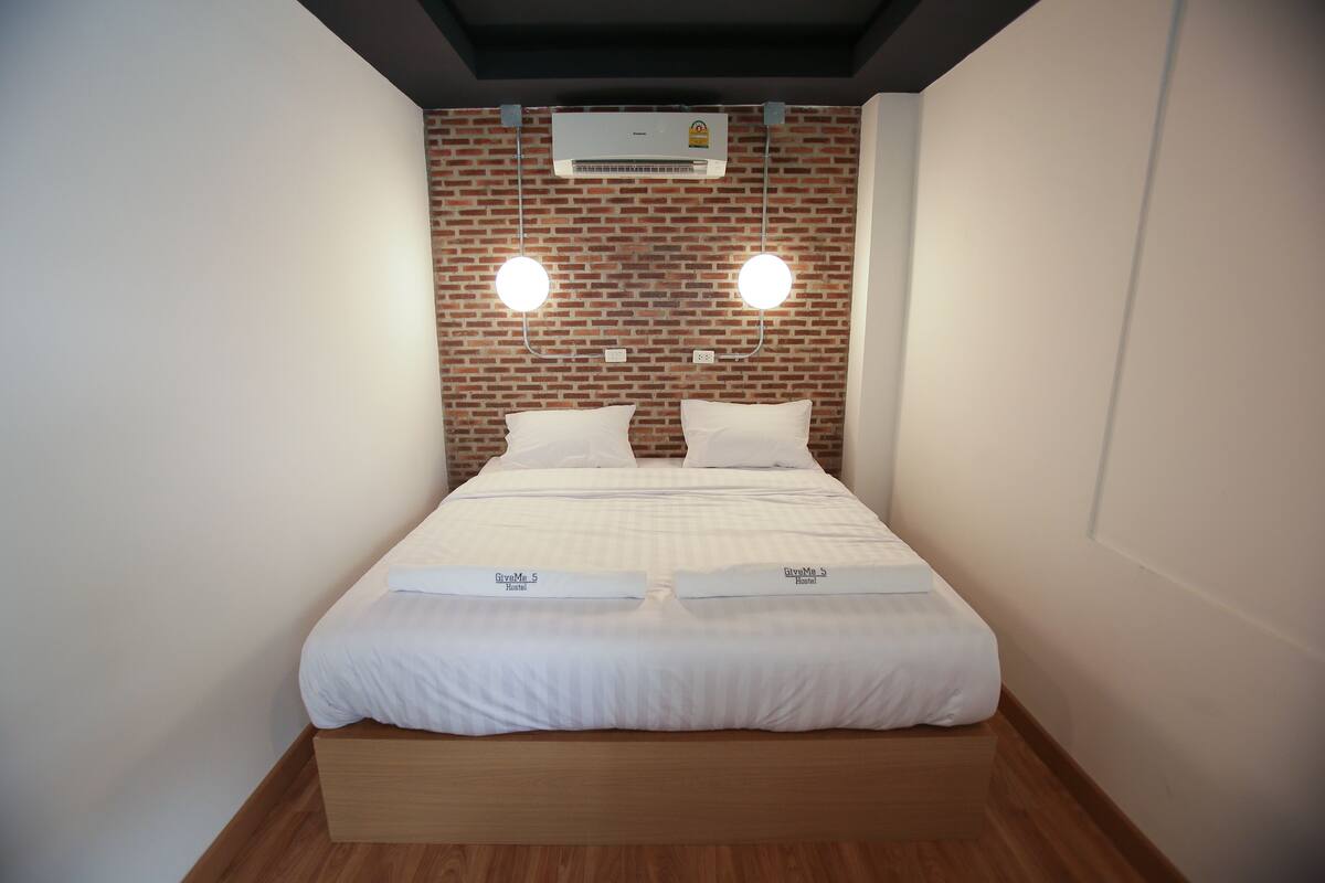 A neatly made bed with white linens and pillows, a brick-patterned wall, and an air conditioning unit above.