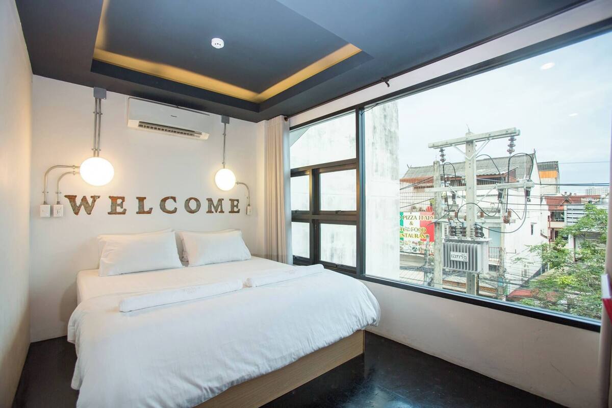 A modern hotel room with a large bed, a "WELCOME" sign, and a view of the cityscape through large windows.