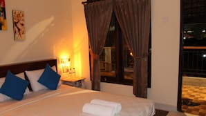 A hotel room with a large bed, blue pillows, a bedside table with a lamp, and a view of the outside. - Chillin Kuta Homestay (Tuban)