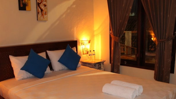 A neatly made bed with white and blue pillows, a rolled white towel, and a nightstand with a lamp and two bottles. - Chillin Kuta Homestay (Tuban)