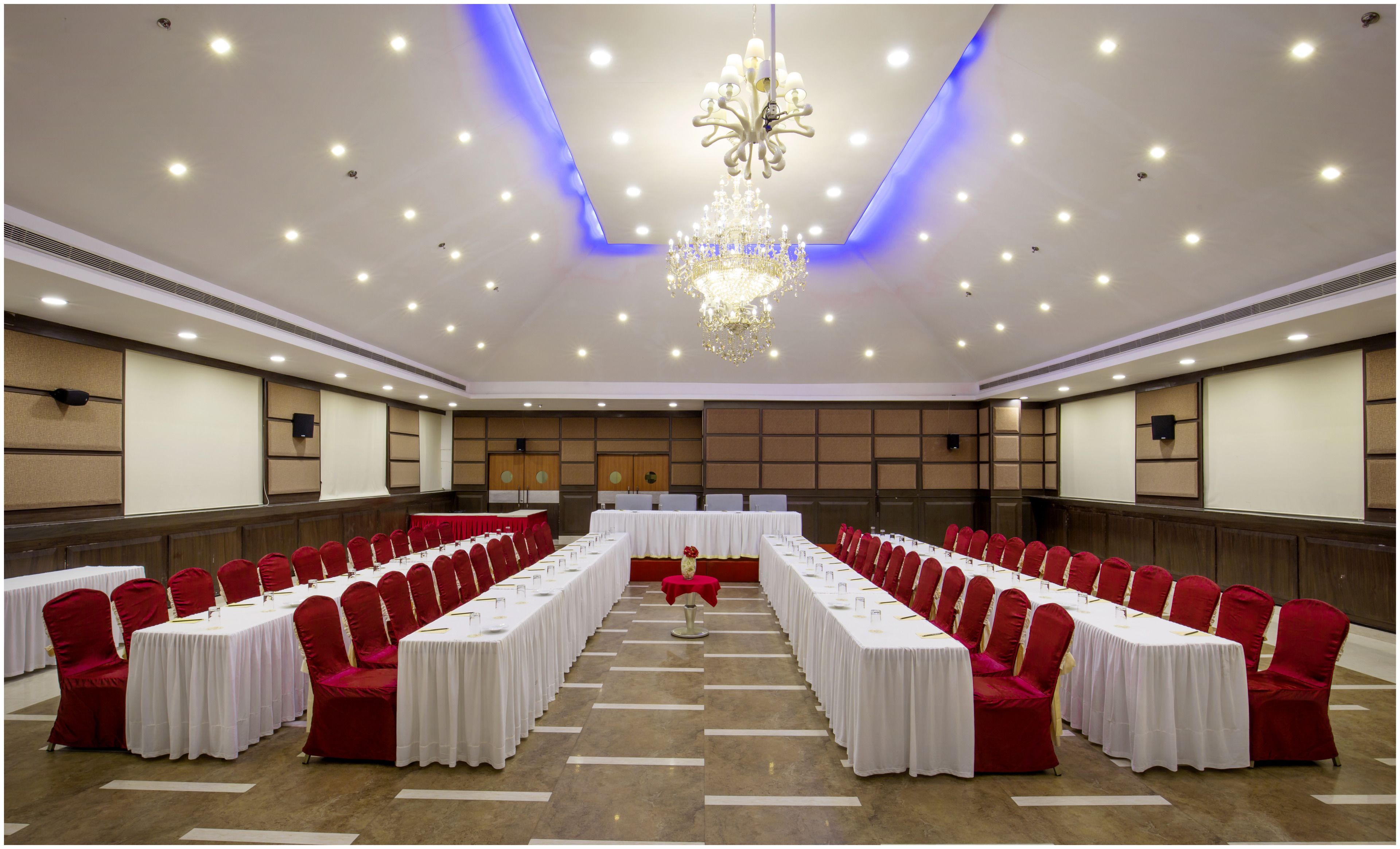 A conference room with long tables and red chairs, equipped with microphones and screens.
