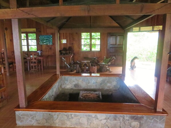 A rustic wooden interior with a stone basin in the center, wooden furniture, and large windows allowing natural light.
