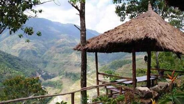 A rustic thatched-roof pavilion with a wooden bench, surrounded by lush greenery and overlooking a valley. - Native Village Inn (Banaue)