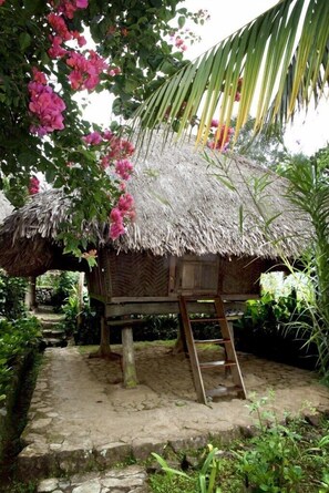 A rustic thatched-roof hut with a wooden ladder leading up to a platform, surrounded by lush greenery and flowering plants.