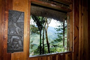 A rustic wooden cabin with a thatched roof and a window overlooking a lush valley.