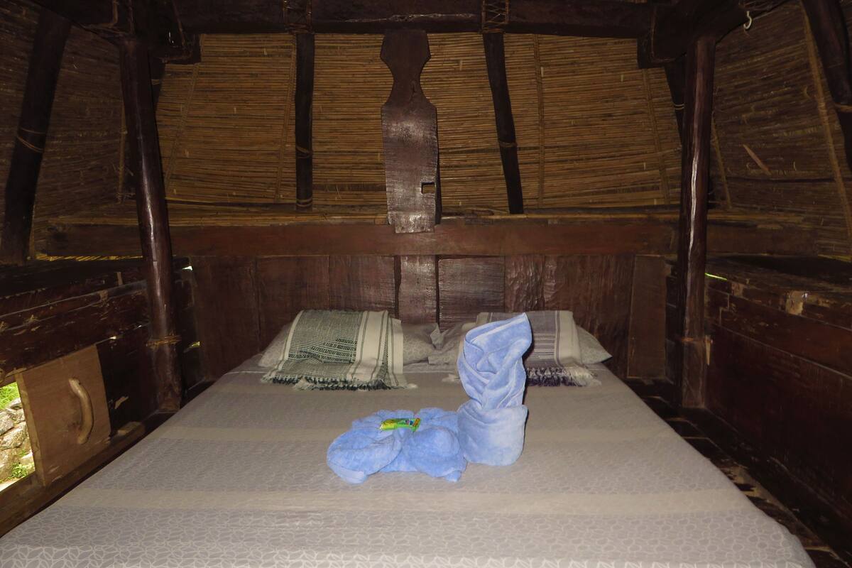 Ein Einzelbett mit gemustertem Matratze, einem gefalteten Handtuch und einer Decke in einem rustikalen Zimmer aus Holz mit Strohdach.