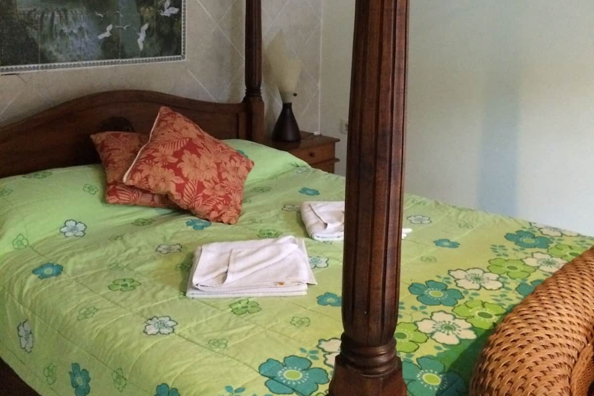 Ein Himmelbett mit einem grün gemusterten Bettbezug, einem hölzernen Kopfteil und einem gewobenen Stuhl.