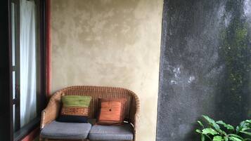 A wicker chair with cushions against a wall, a window with curtains, and a plant beside the wall.