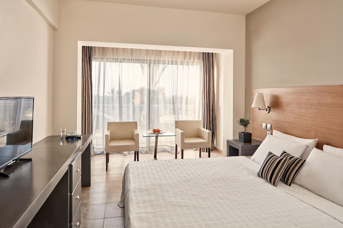 Une chambre d’hôtel avec un lit, un bureau avec un ordinateur, deux chaises, une petite table avec un verre d’eau et une fenêtre avec des rideaux.