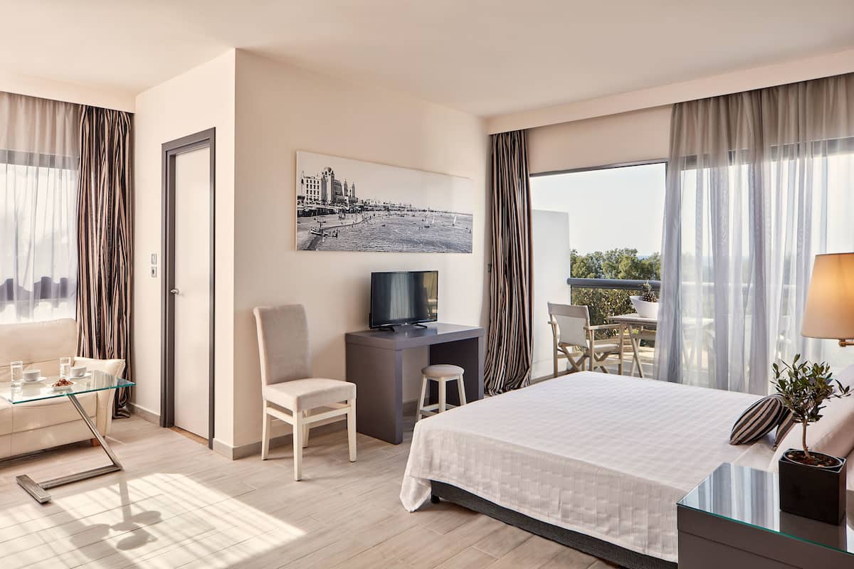 Une chambre d’hôtel moderne équipée d’un lit, d’un bureau avec une télévision, d’une table en verre et d’un balcon avec vue.