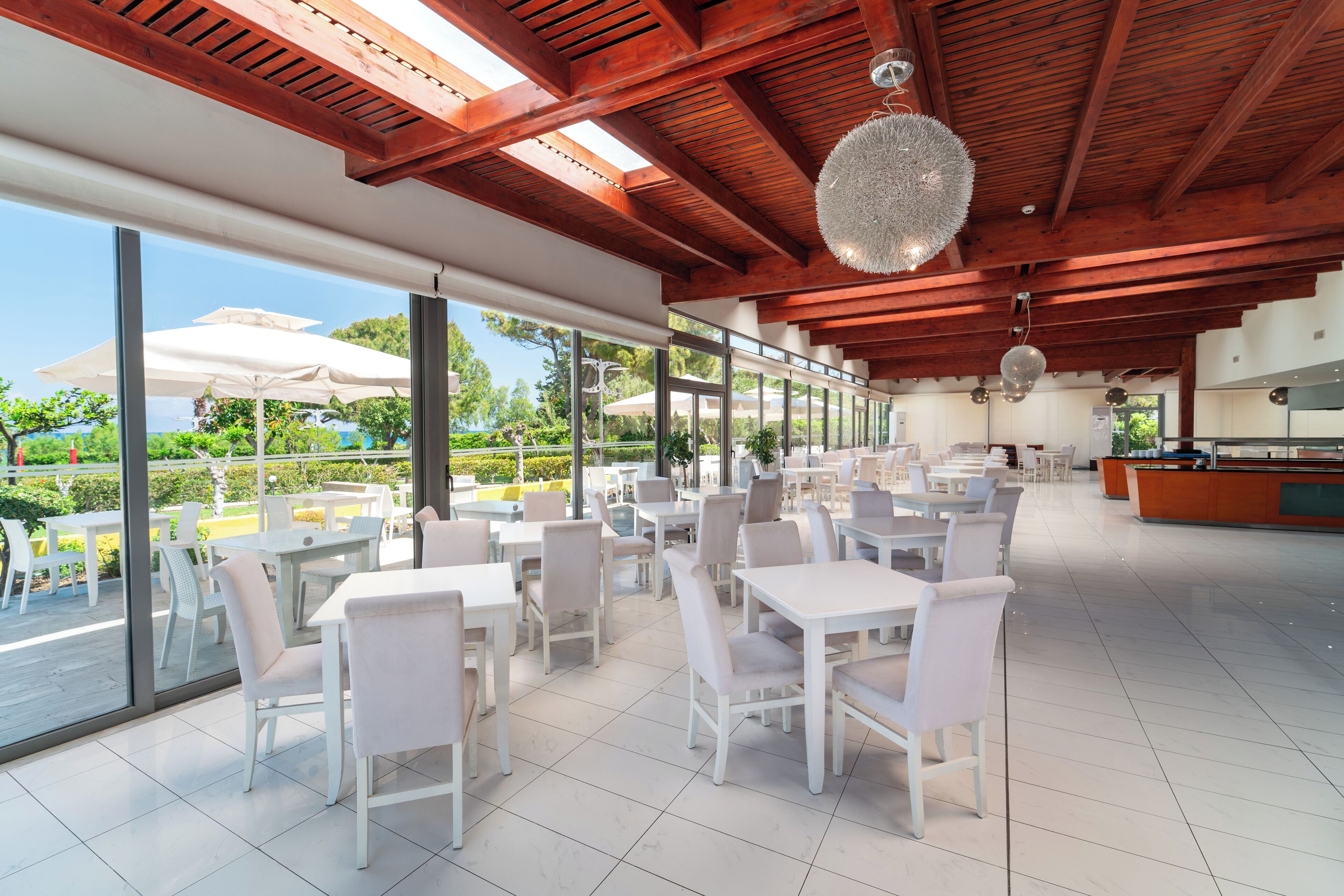 A spacious outdoor dining area with white tables and chairs, large windows, and a wooden ceiling.