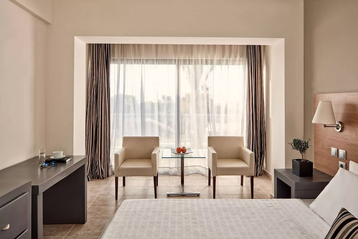 Une chambre d’hôtel comprenant un lit, deux fauteuils, une petite table, un bureau, une lampe et une vue sur la ville grâce à de grandes fenêtres.