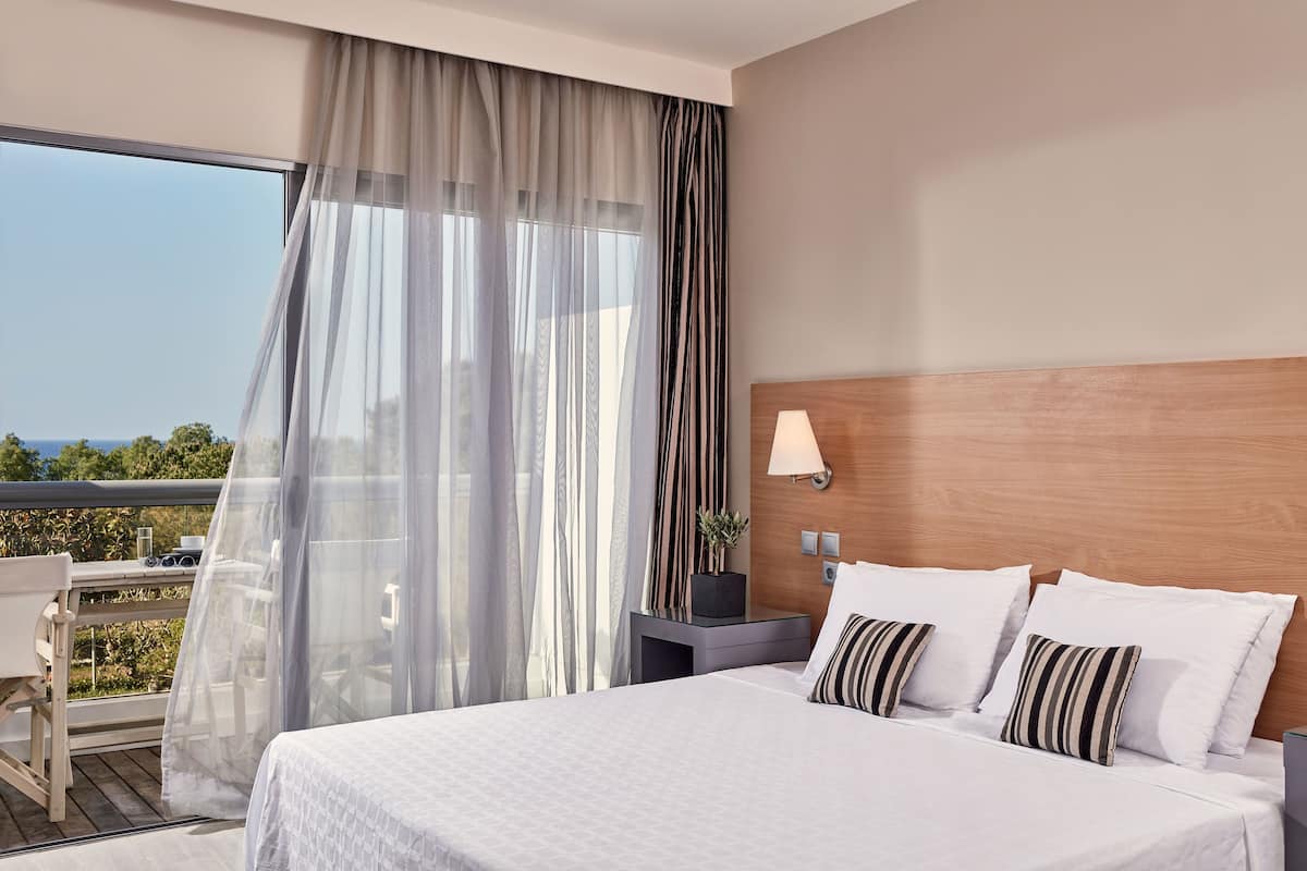 Une chambre d’hôtel avec un grand lit, une table de chevet avec une lampe, des rideaux légers et une vue sur l’extérieur.