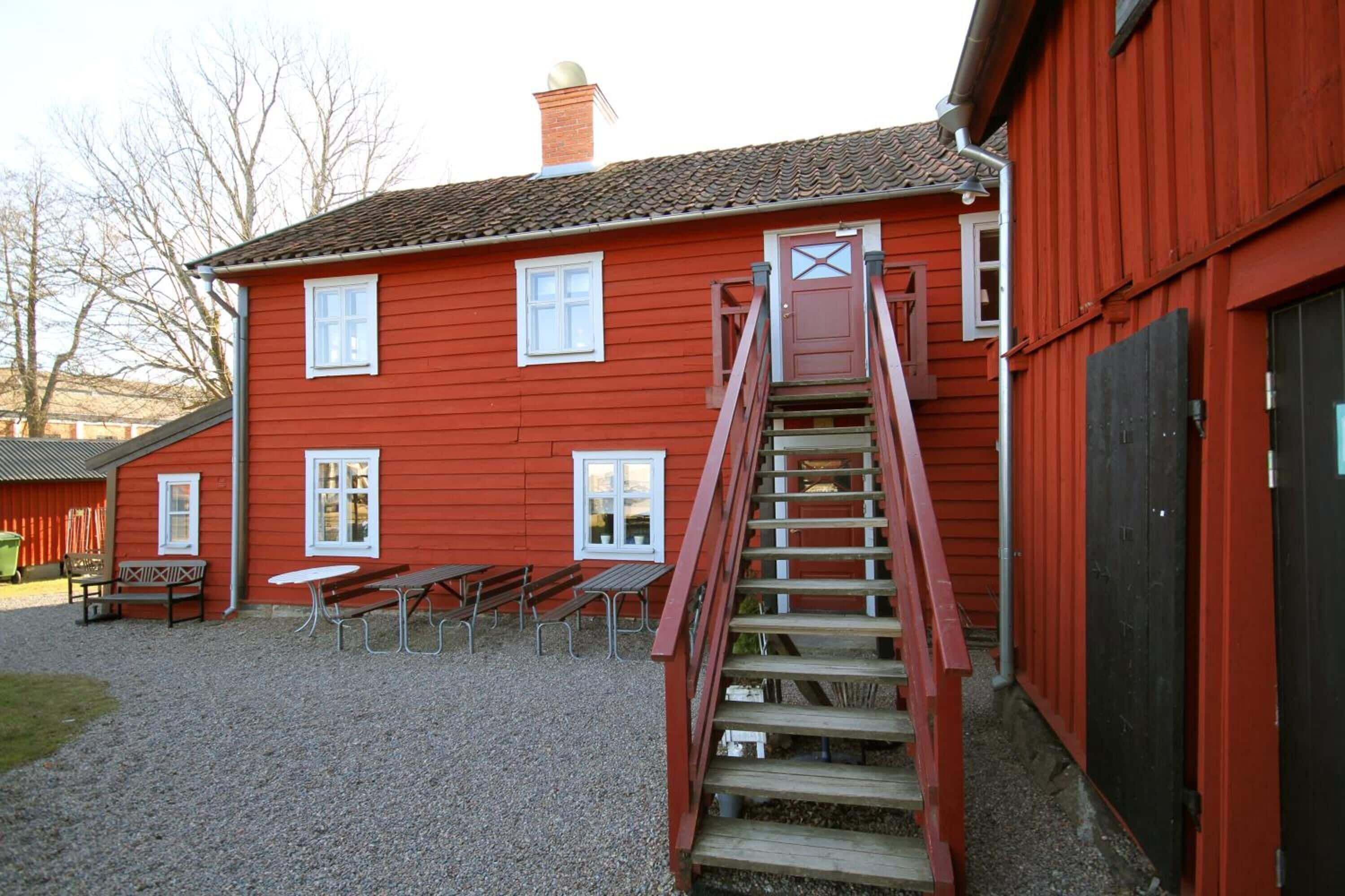 A red wooden building with a staircase leading to a door, outdoor seating, and a gravel ground.