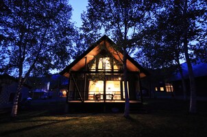 A small, cozy cabin with a sloped roof, surrounded by trees, and a lit interior visible through the windows.