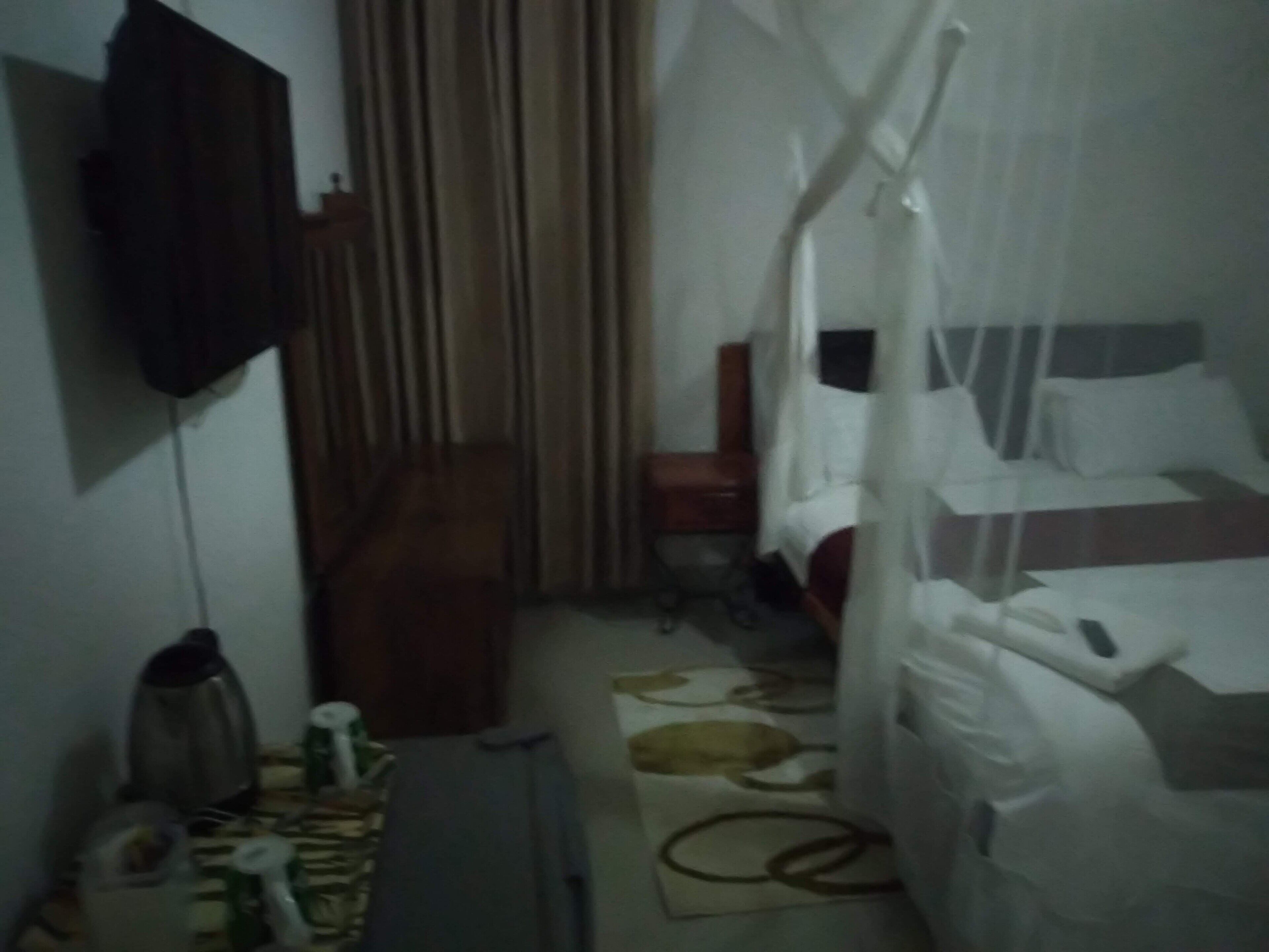 Una habitación de hotel tenuemente iluminada, con una cama, una mesita pequeña, una silla y un televisor montado en la pared.