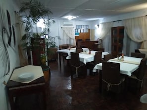 A dining area with tables set for four, a large potted plant, and a reception desk in the background. - Kuku Royal Lodge (Ndola)