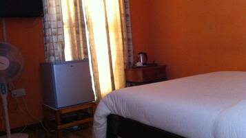 Quarto com cama, um ventilador, uma mesinha com chaleira e televisão em móvel.