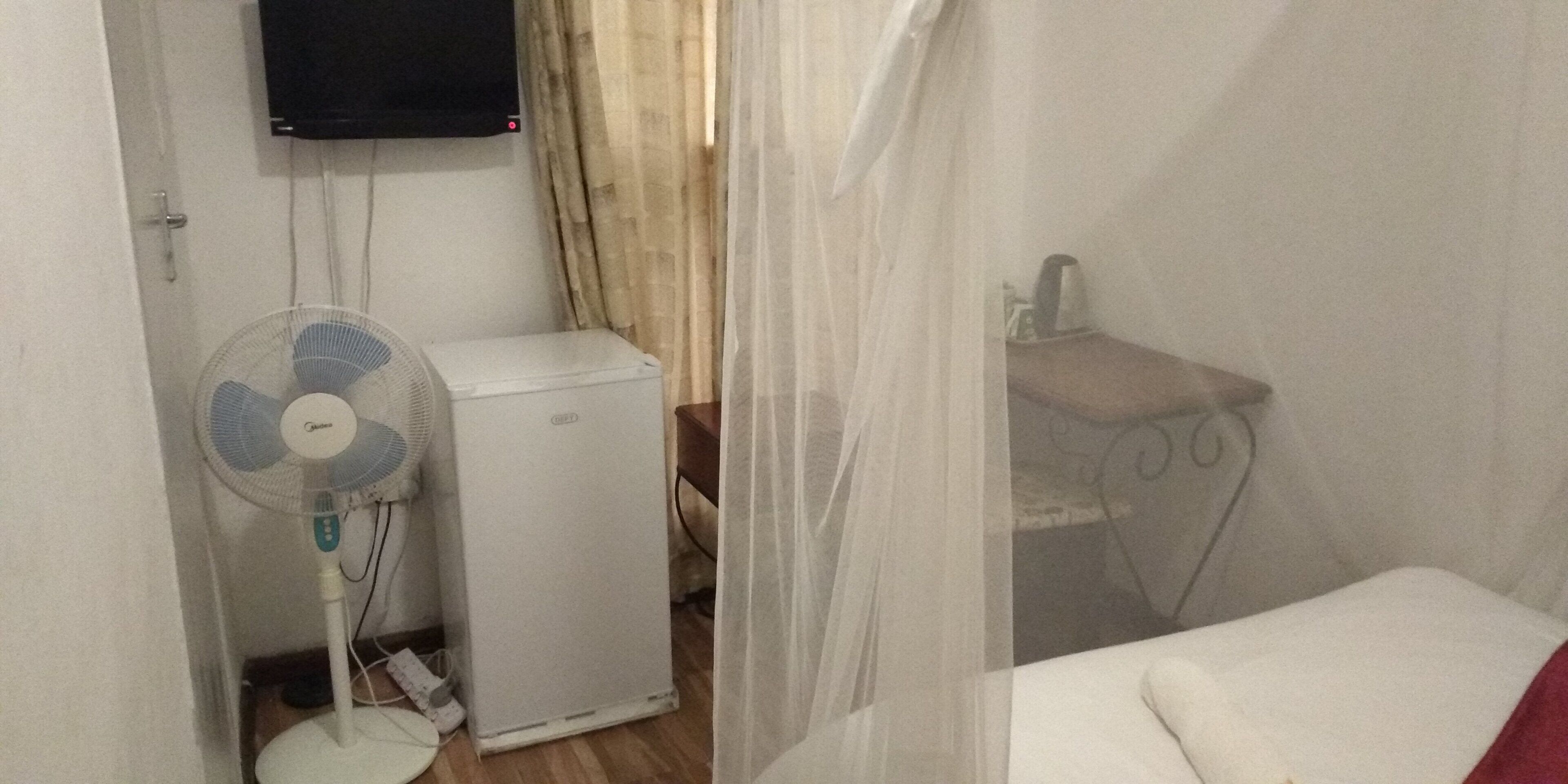 Una habitación con una cama, una mesita de noche, un refrigerador blanco pequeño, un ventilador de pie y un televisor montado en la pared.
