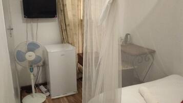 Una habitación con una cama, una mesita de noche, un refrigerador blanco pequeño, un ventilador de pie y un televisor montado en la pared.