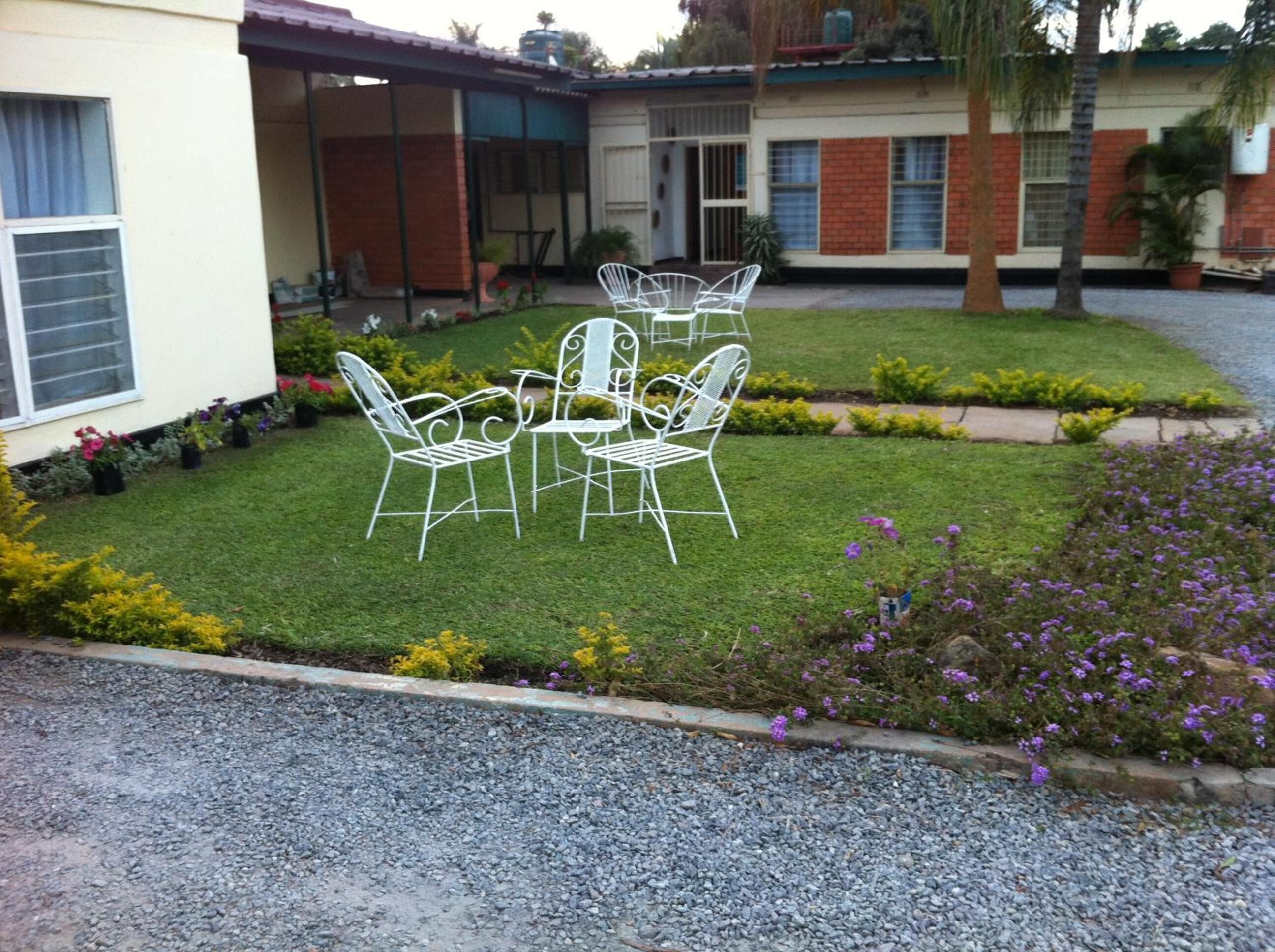 A courtyard with a lawn, white metal chairs, and a small garden with flowers.