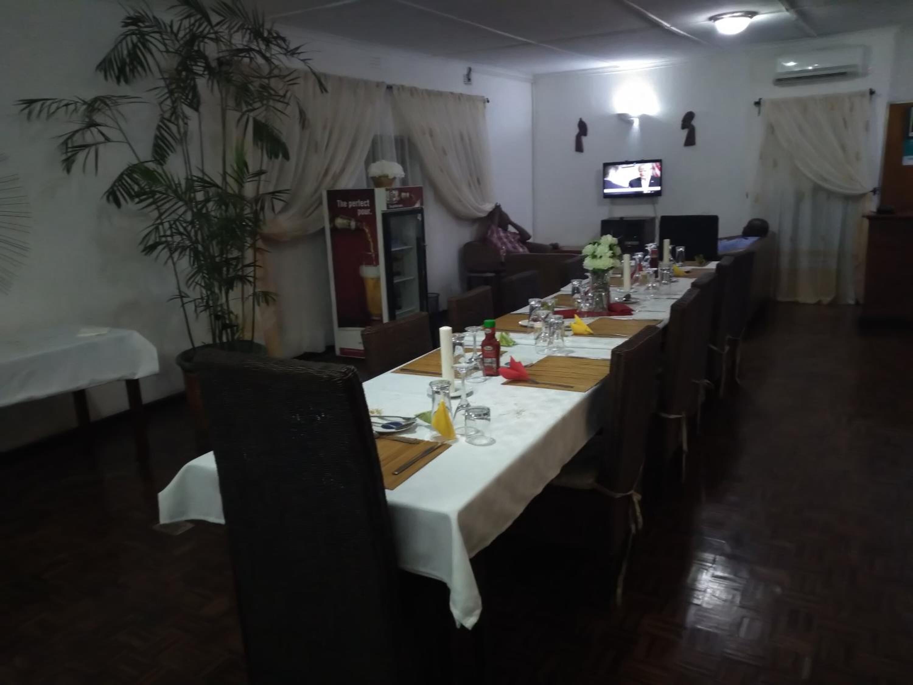 Un comedor con una mesa larga dispuesta para una comida, sillas, un televisor en la pared y una planta en maceta.