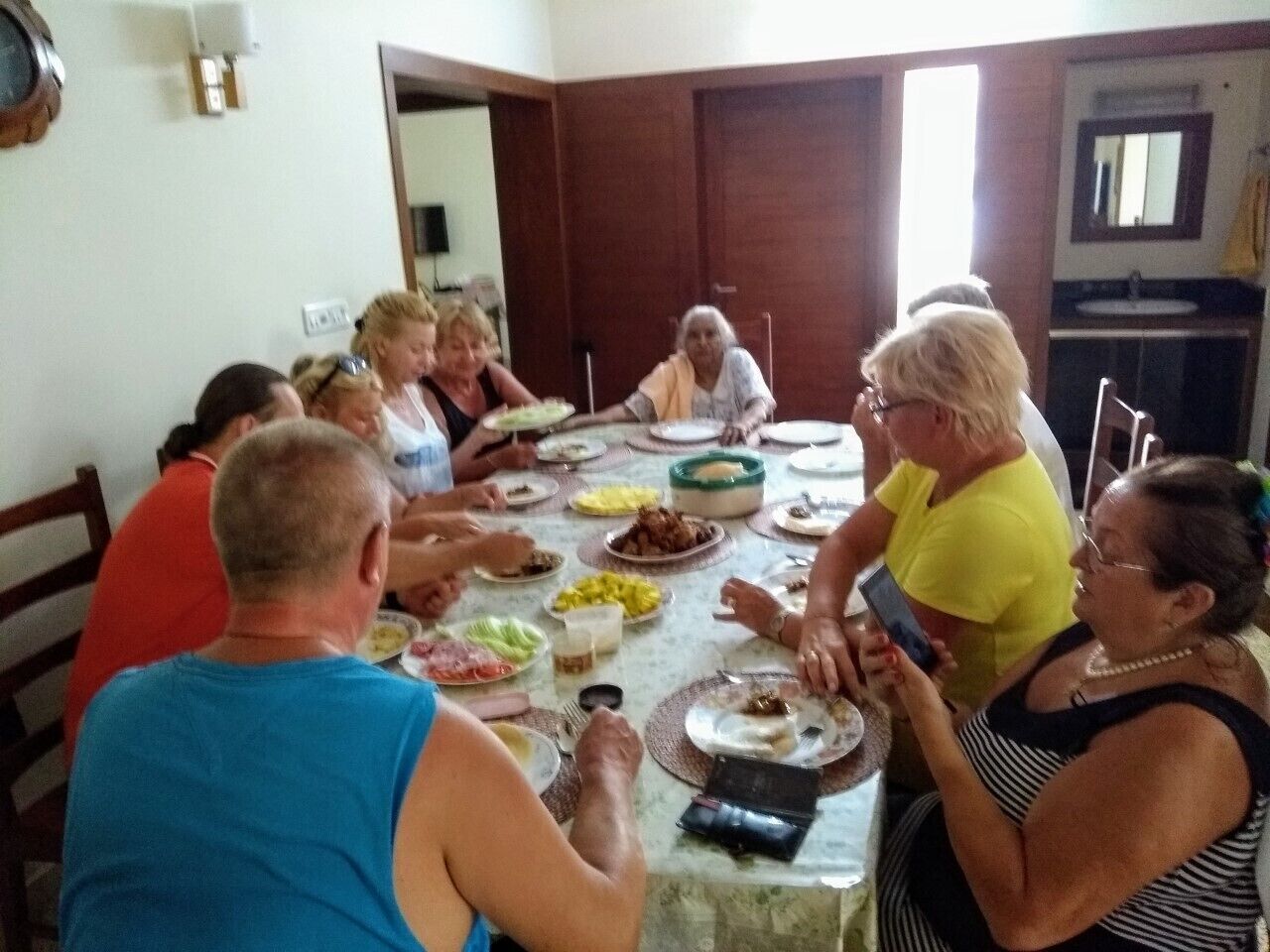 A group of people seated around a table with various dishes, in a room with wooden doors and a mirror on the wall.