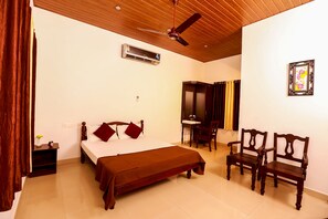 A hotel room with a bed, two wooden chairs, a desk, an air conditioning unit, and a ceiling fan. - Baywatch Beach Resort (Ambalapuzha)