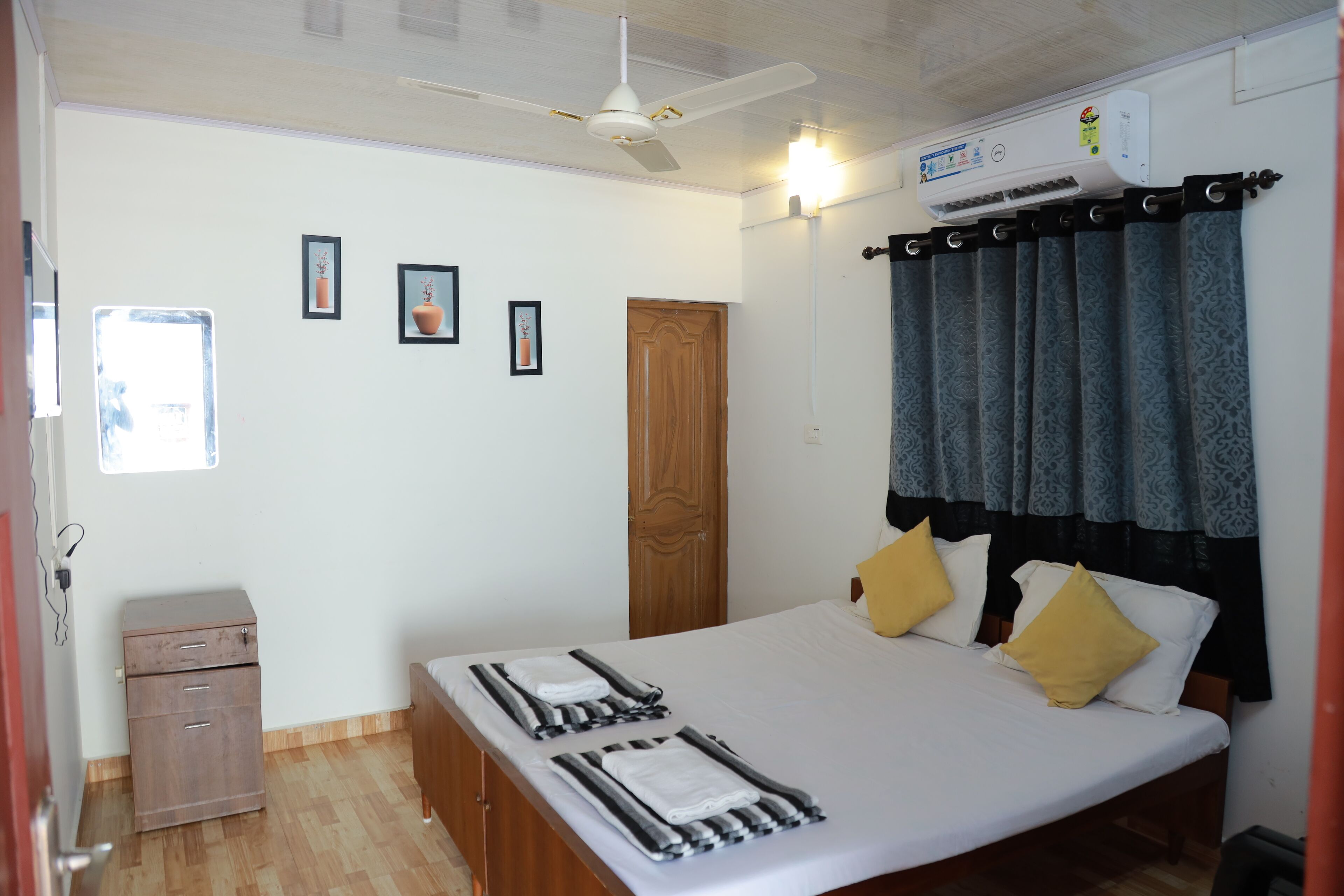 A bedroom with a bed, a ceiling fan, an air conditioning unit, a wooden wardrobe, and a window.