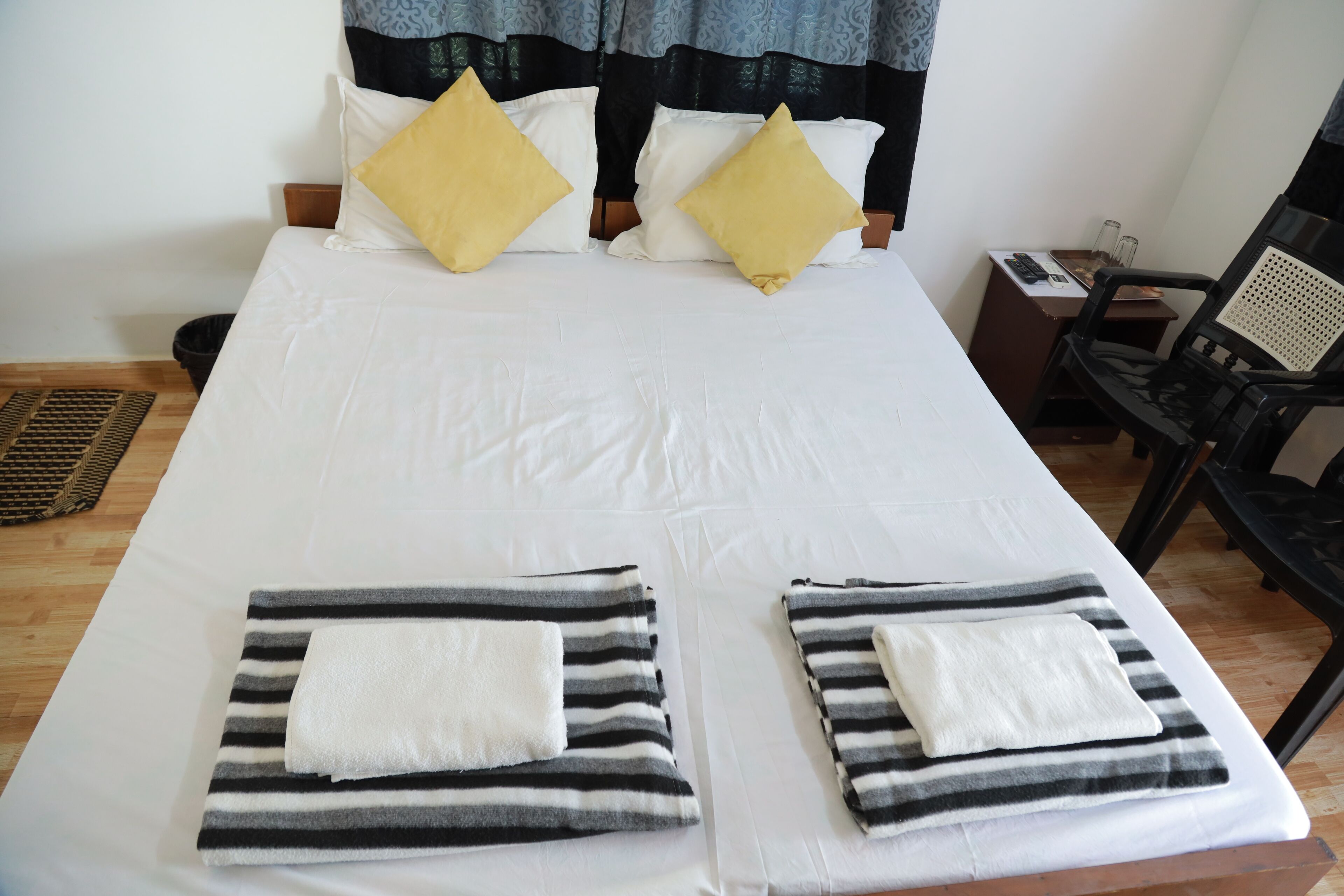 A double bed with white and yellow pillows, two striped towels, a wooden headboard, a nightstand with a phone and glasses, and a black chair.