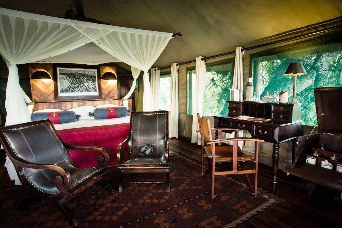 A room with a bed, two leather chairs, a desk, and a large window with a view of greenery.