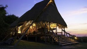 A thatched-roof structure with wooden stairs leading to a platform, surrounded by vegetation. - Selinda Camp (Selinda Reserve)