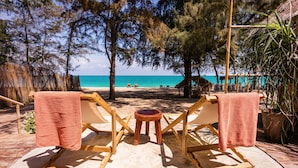 Dos sillones de madera con toallas, una mesita redonda y vista a la playa con árboles y una estructura de techo de paja al fondo.
