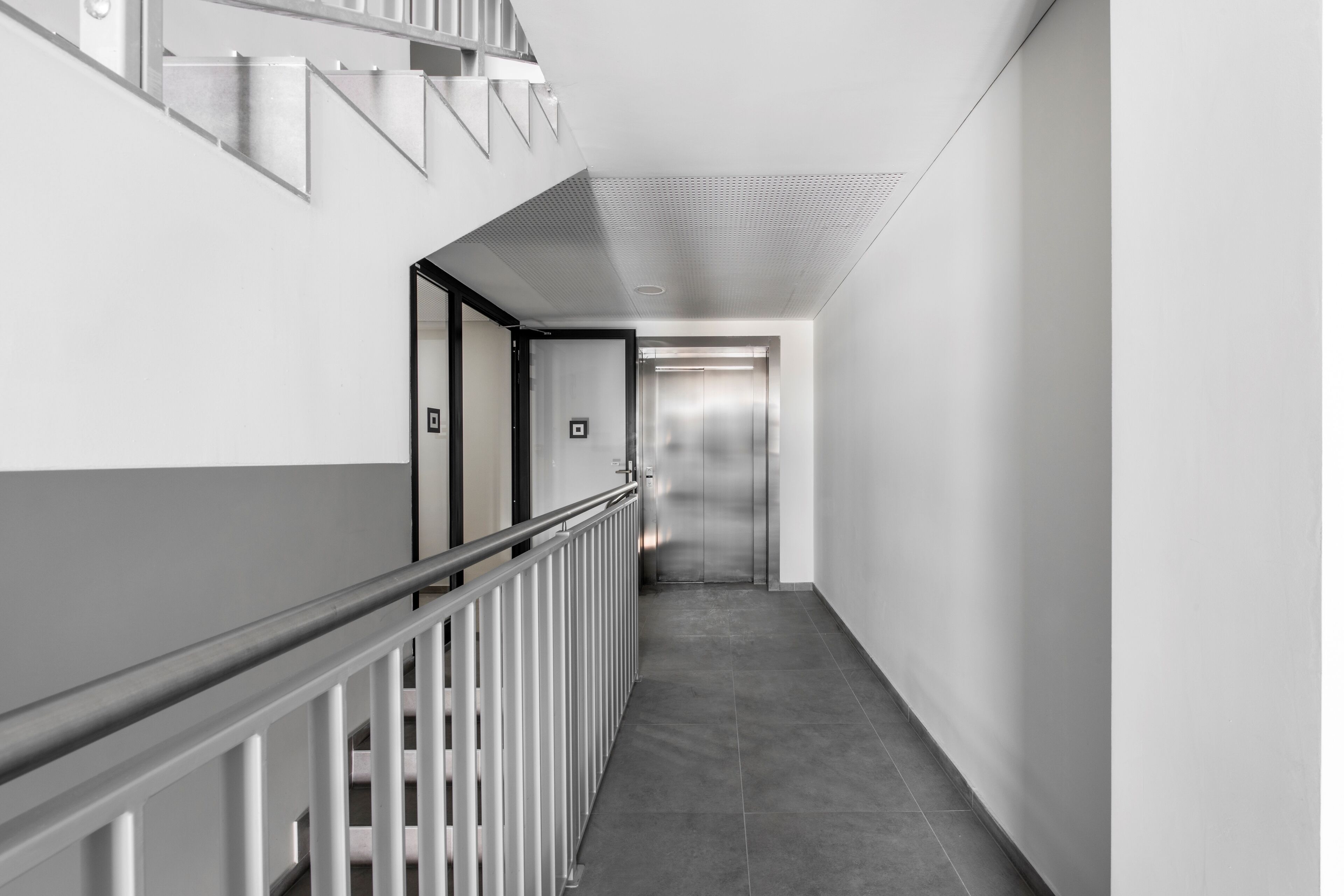 A modern, clean hallway with a metal railing, a staircase, and an elevator.