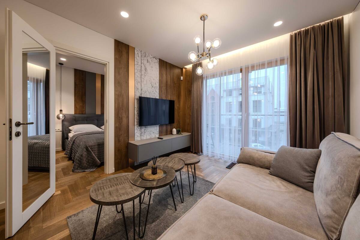 A modern living room with a sofa, a TV mounted on the wall, a dining area with a table and chairs, and a large window with curtains.