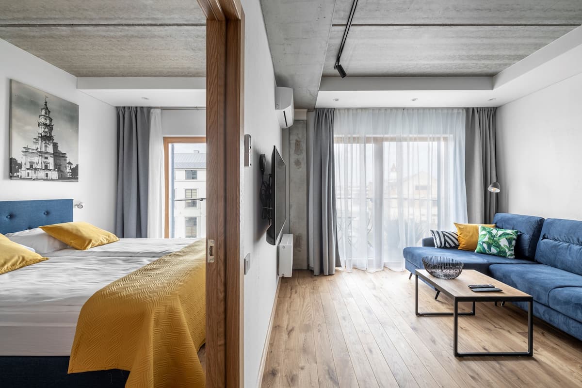 A modern hotel room with a bed, a sofa, a wooden coffee table, and a view of the city through large windows.