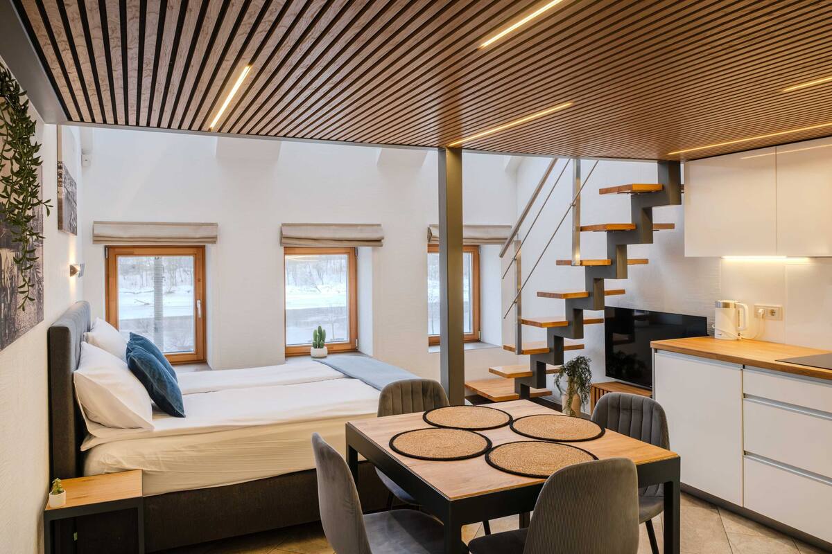 A modern interior with a bed, dining table, chairs, kitchen area, and a staircase.