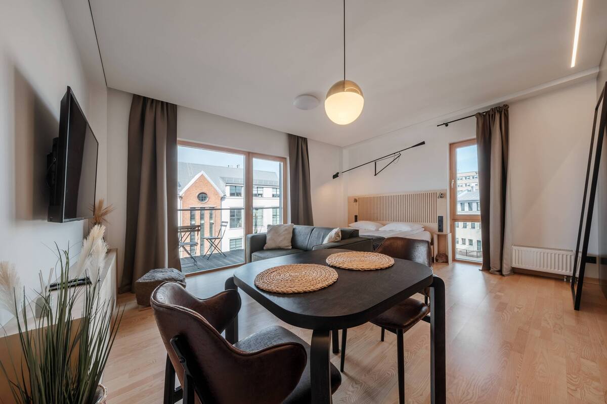 A modern living room with a dining area, large windows, a flat-screen TV, and a view of the cityscape.
