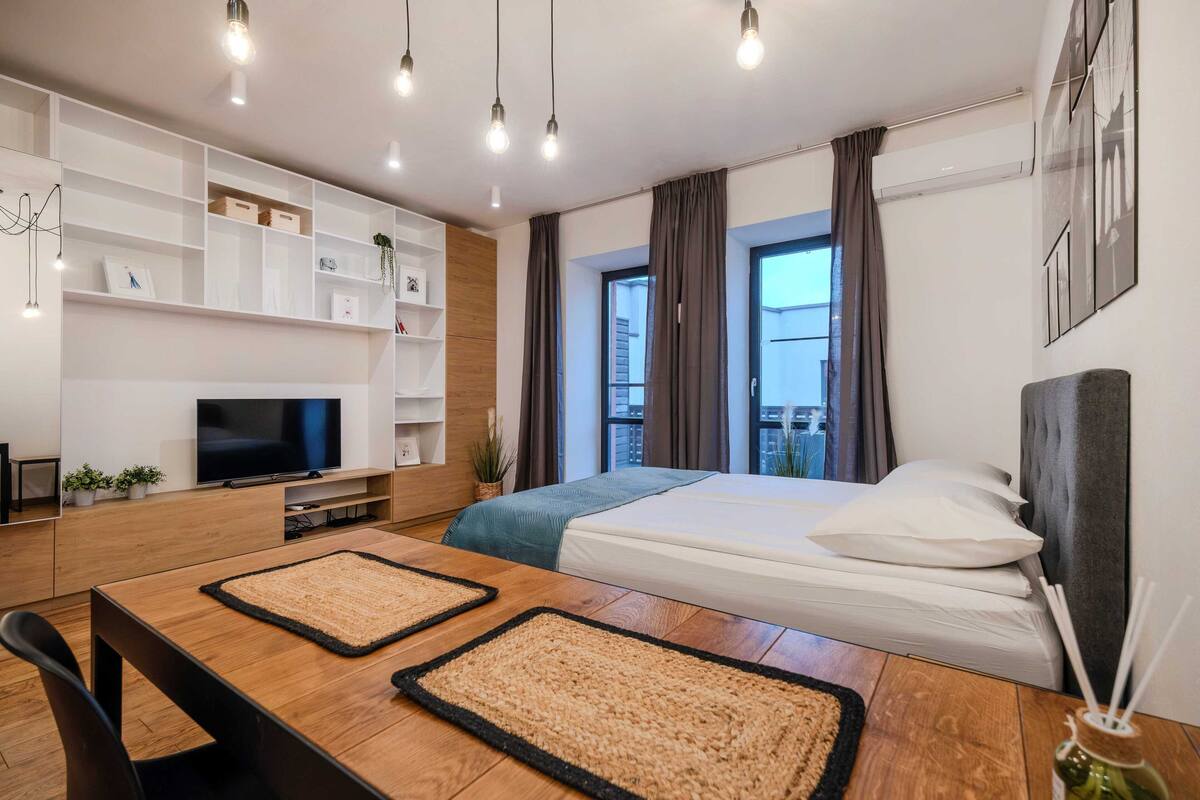 A modern hotel room with a bed, a TV, a wooden desk, and a large window with curtains.