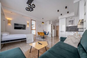 A modern, well-lit living room with a sofa, a small table, and a bed. There is a dining area with chairs and a table, and a kitchen with cabinets and a sink. - King Mindaugas Apartments (Kaunas)