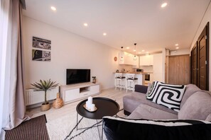 A modern living room with a sofa, a coffee table, a TV, and a kitchen area in the background. - King Mindaugas Apartments (Kaunas)