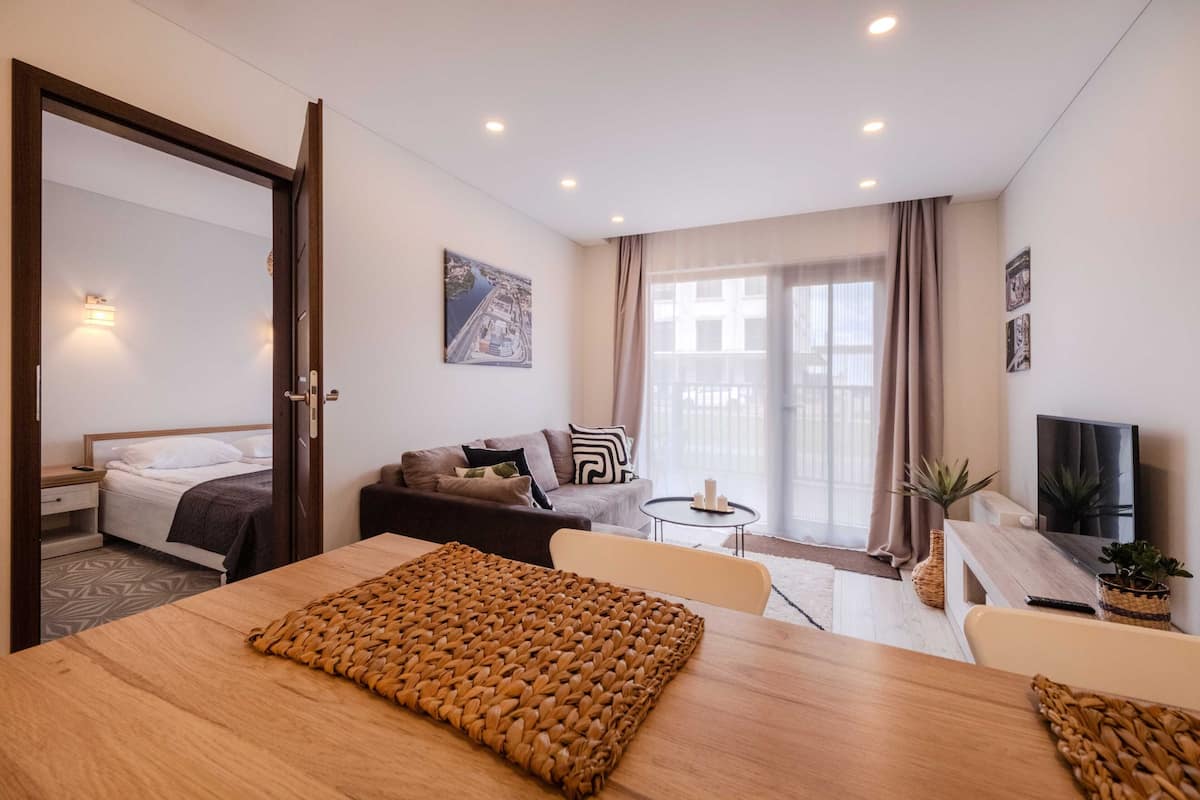 A hotel room with a bed, a sofa, a dining table, a TV, and a balcony with a view.