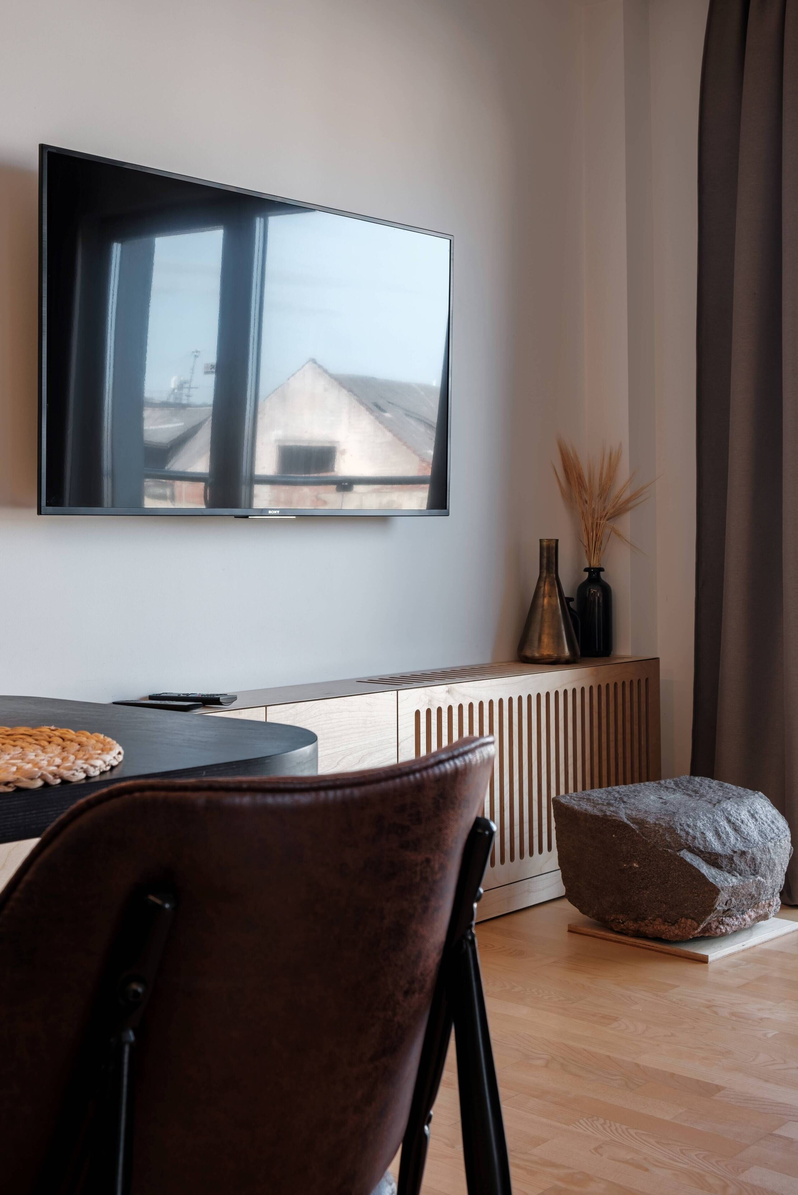 Una sala moderna con un televisor de pantalla plana grande montado en la pared, una mesa de comedor de madera con sillas, un radiador debajo y una gran escultura de roca en el suelo.