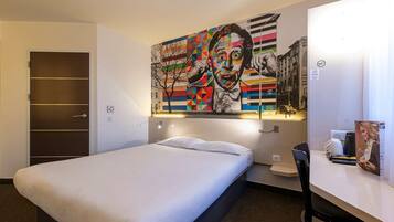 A hotel room with a bed, a desk with a chair, a wall-mounted artwork, and a door with a shelf.