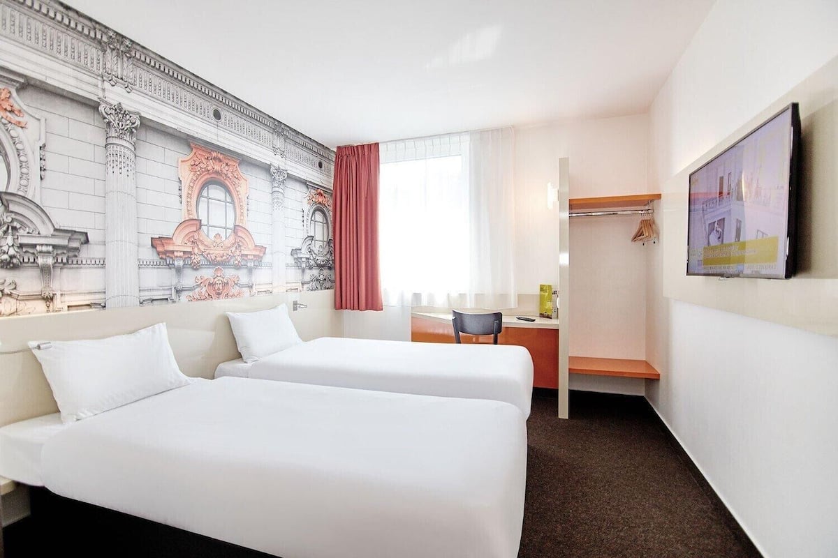 Quarto de hotel com duas camas, uma escrivaninha, uma cadeira, uma televisão e um guarda-roupa.