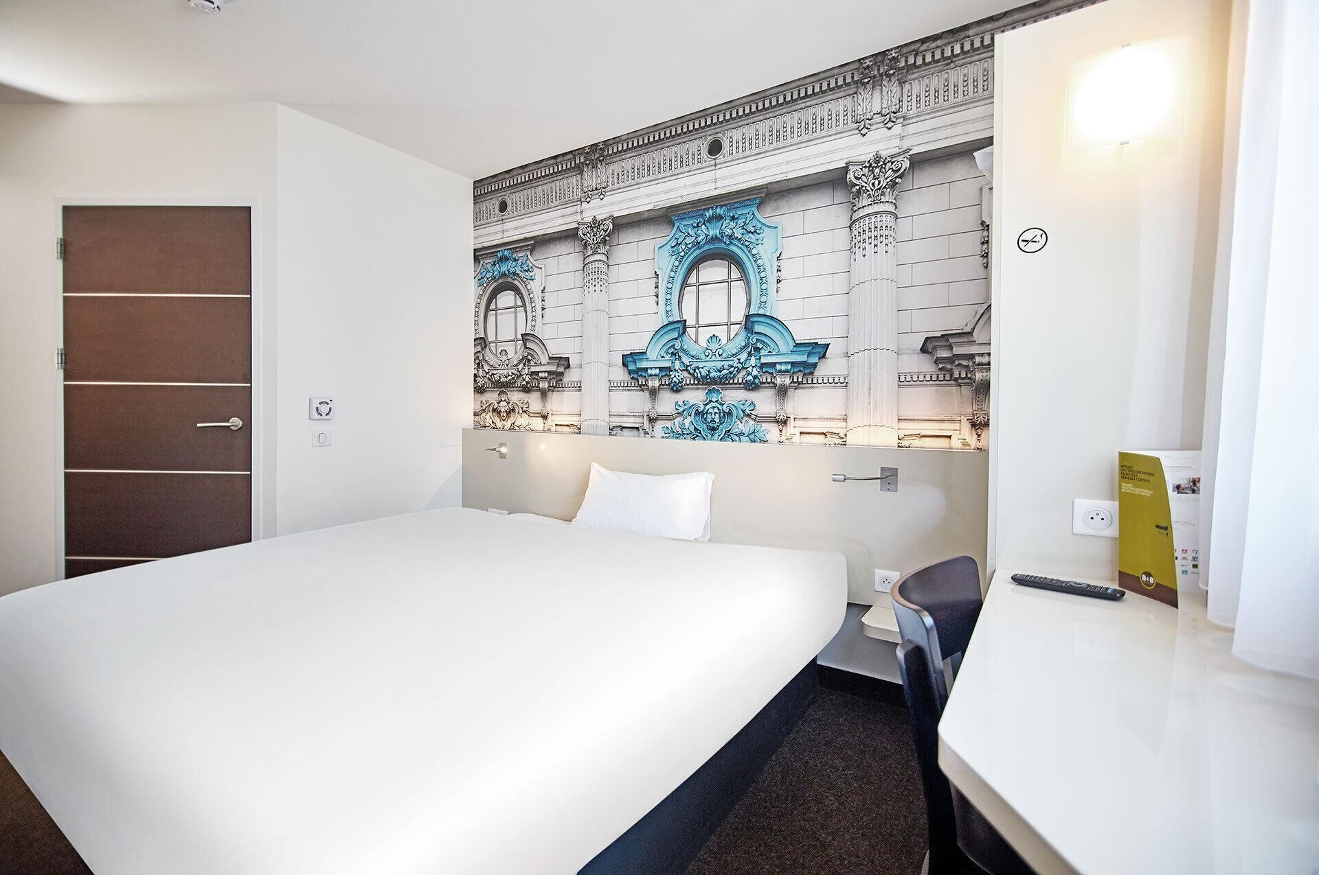 A hotel room with a large bed, a desk, a chair, and a decorative wall with architectural elements.