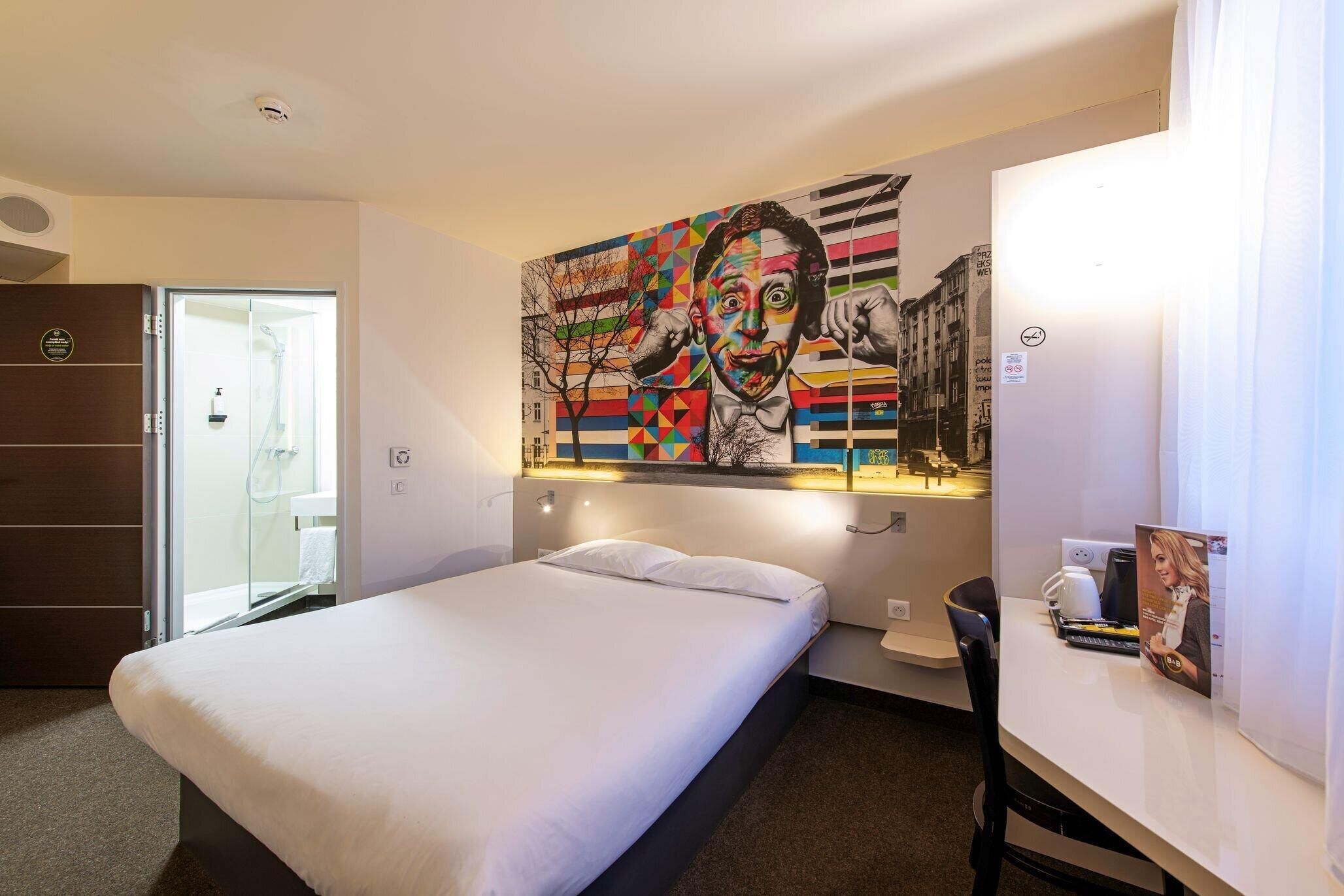 A hotel room with a bed, a shower, a desk, and a large colorful mural on the wall.
