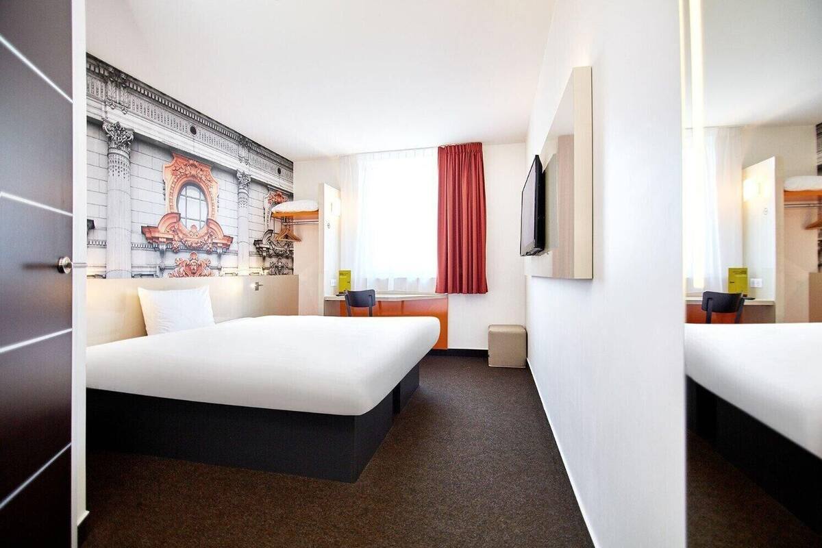 Quarto de hotel com uma cama grande, uma escrivaninha, uma cadeira, uma televisão e um mural na parede.